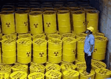 a man in a hard hat is standing in front of a pile of yellow barrels with radioactive symbols on them .