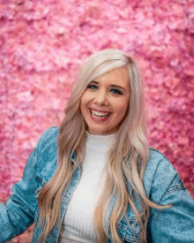 the woman is wearing a denim jacket and smiling in front of a pink wall of flowers .