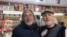 two men are posing for a picture in front of a store . one of the men has long hair and a beard .