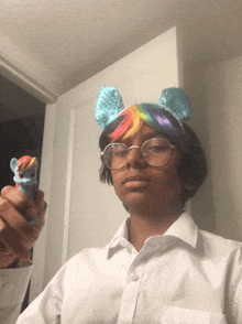 a boy wearing a rainbow colored wig and glasses holds a toy pony