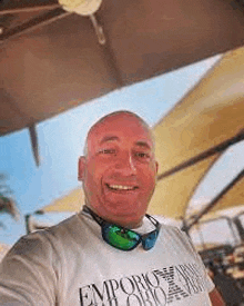 a bald man wearing sunglasses and a t-shirt is taking a selfie on the beach .