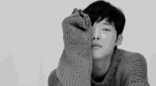 a black and white photo of a young man in a sweater covering his face with his hand .