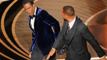 two men in suits are dancing on a stage and one is wearing a blue jacket