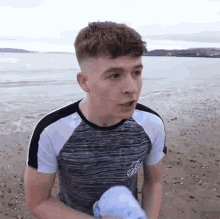 a young man standing on a beach wearing a grey and white shirt with the letter b on the sleeve