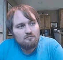 a man with a beard and a blue shirt is sitting in a kitchen looking at the camera .