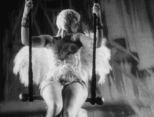 a black and white photo of a woman in a feathered dress sitting on a swing .