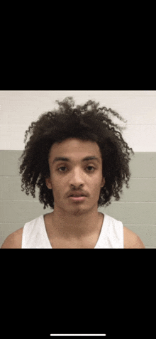 a young man with curly hair and a white tank top looks at the camera