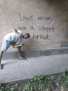 a young man stands in front of a wall that says " look mum i am a street artist "