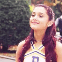 ariana grande is wearing a cheerleading uniform and smiling while standing in front of a bush .