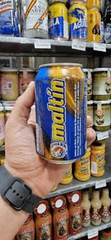 a person is holding a can of maltin beer in their hand in front of a shelf .