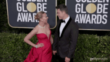 a woman in a red dress and a man in a tuxedo holding hands in front of a sign that says golden globe