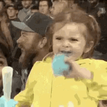 a little girl in a yellow jacket is eating a blue ice cream cone in front of a crowd .