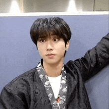 a young man is standing in front of a blue wall wearing a black jacket and a floral shirt .