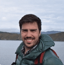 a man wearing a green jacket and a backpack is smiling in front of a body of water .