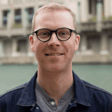 a man wearing glasses and a denim jacket smiles