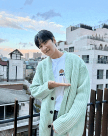 a man wearing a green cardigan and a white t-shirt is standing on a balcony with a building in the background