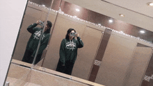 a woman taking a picture of herself in a bathroom mirror wearing a green sweatshirt that says masters on it