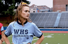 a woman wearing a whs yellowjackets shirt stands on a field with her hands on her hips