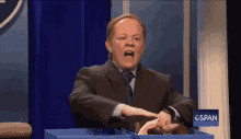 a man in a suit and tie is standing in front of a blue curtain and holding something in his hand .