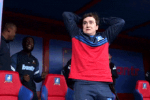 a man in a red and blue shirt is standing in a soccer dugout with his hands on his head .