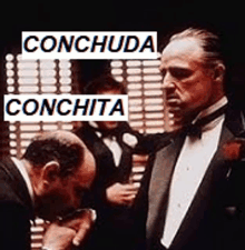 a man in a tuxedo is kissing another man 's hand with the words conchuda conchita written above him .