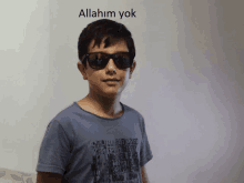 a boy wearing sunglasses stands in front of a wall that says allahum yok