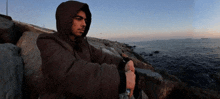 a man in a hooded jacket is sitting on a rock near the water