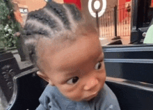 a baby with braids on his head is sitting in a shopping cart and looking at the camera .