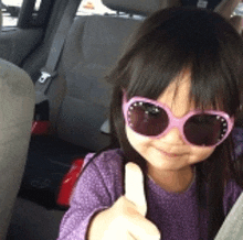 a little girl wearing sunglasses and a purple shirt is giving a thumbs up