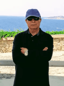 a man wearing a blue hat and sunglasses stands with his arms crossed in front of the ocean