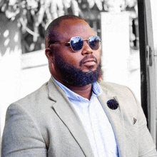 a man with a beard wearing sunglasses and a suit is standing next to a window .