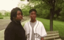 a couple of men standing next to each other in a park . one of the men is wearing a baseball jersey .