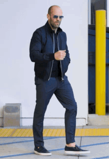 a bald man wearing sunglasses and a black jacket is walking down a sidewalk