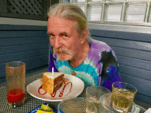 a man in a tie dye shirt sits at a table with a piece of cake