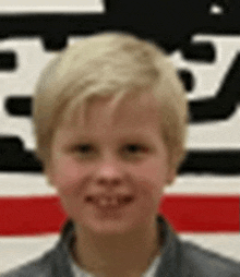 a young boy with blonde hair is smiling in front of a red , white and black sign .