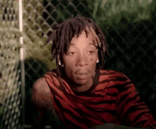 a young man with dreadlocks is sitting in front of a chain link fence wearing a zebra print shirt .