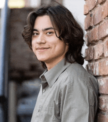 a young man leans against a brick wall and smiles for the camera