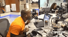 a man in an orange shirt is sitting at a desk in front of a laptop and a microphone with the breakfast club written on the table