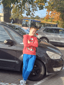 a boy leans against a black car with a bus in the background that says ' tk ' on it