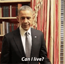 a man in a suit and tie is standing in front of a bookshelf and asking if he can live .