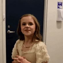 a little girl stands in front of a blue door with a hand sanitizer dispenser hanging on the wall behind her