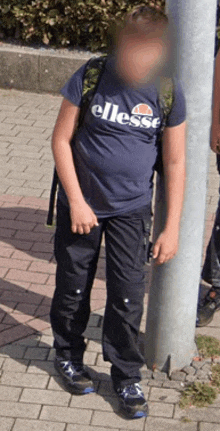 a boy wearing a blue ellesse t-shirt leans against a pole