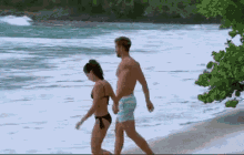 a man and a woman in bikinis are walking on the beach holding hands .