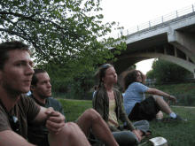 a group of people sitting on the grass under a bridge