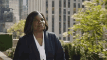 a woman in a suit and white shirt is standing on a balcony with a city in the background .