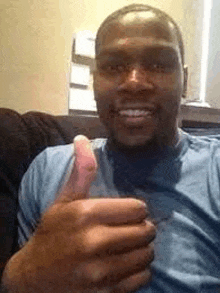 a man in a blue shirt is sitting on a couch giving a thumbs up .