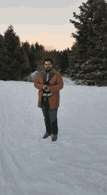 a man in a brown coat stands in the snow with trees in the background