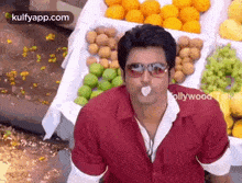 a man wearing sunglasses is sitting in front of a fruit stand with a piece of gum in his mouth .