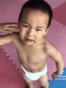 a baby in a diaper is standing on a pink mat and making a funny face .