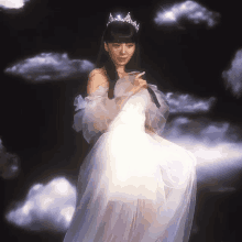 a woman in a white dress and tiara stands in front of a cloudy sky